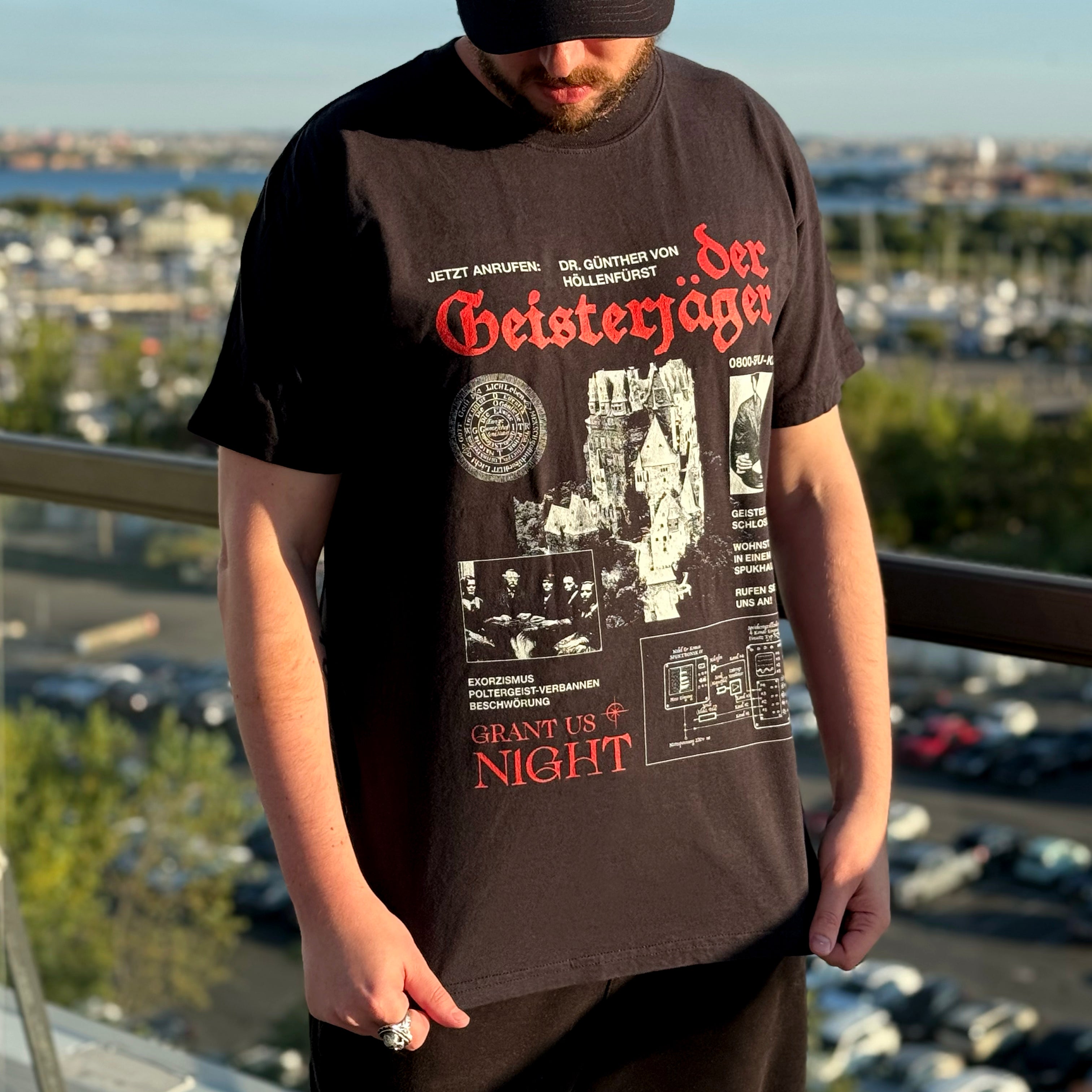 Model wearing Grant Us Night Geisterjager black t-shirt with castle imagery and old photography and graphic design and German text on a blurred castle background hanging outdoors