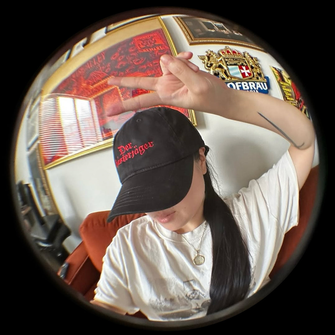 Person wearing a Grant Us Night Geisterjager black baseball cap with 'Der Geisterjager' red text embroidered against a  black and white castle background in an indoor setting with decorative elements.