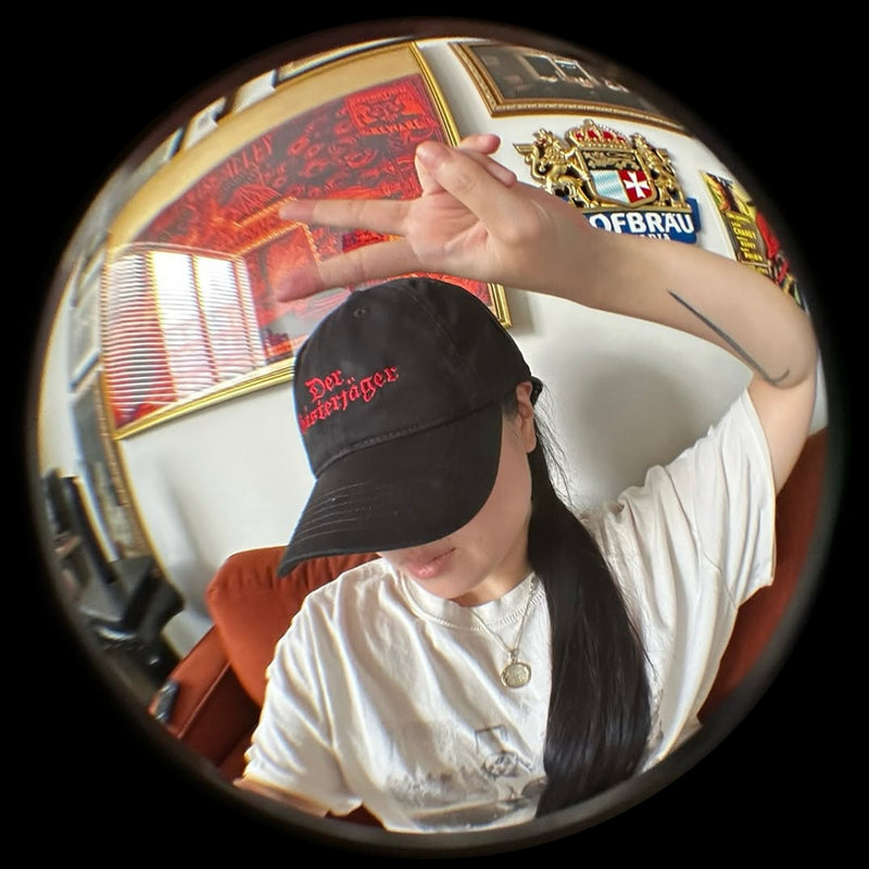 Person wearing a Grant Us Night Geisterjager black baseball cap with 'Der Geisterjager' red text embroidered against a  black and white castle background in an indoor setting with decorative elements.