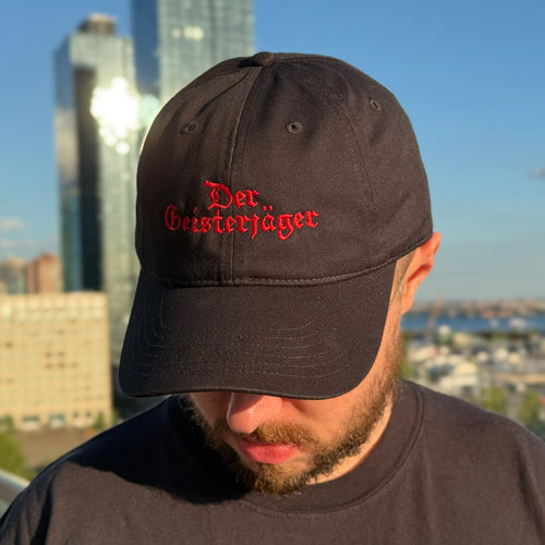 Man wearing a Grant Us Night Geisterjager black baseball cap with 'Der Geisterjager' red text embroidered against a  black and white castle background against a cityscape background