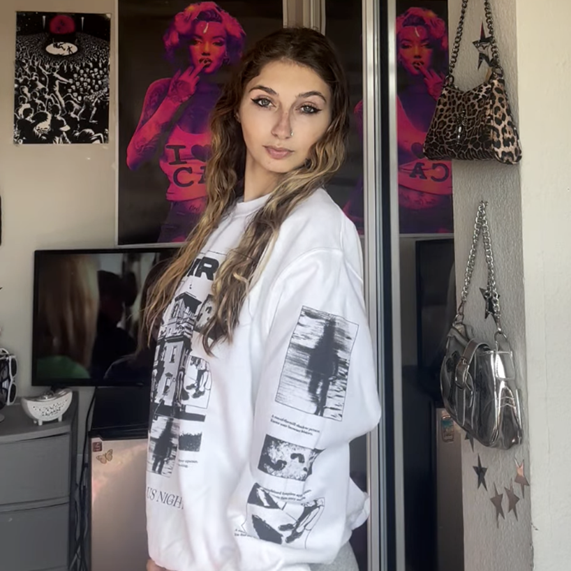 Woman wearing a white Grant Us Night sweatshirt with black graphics in a room with posters and bags.