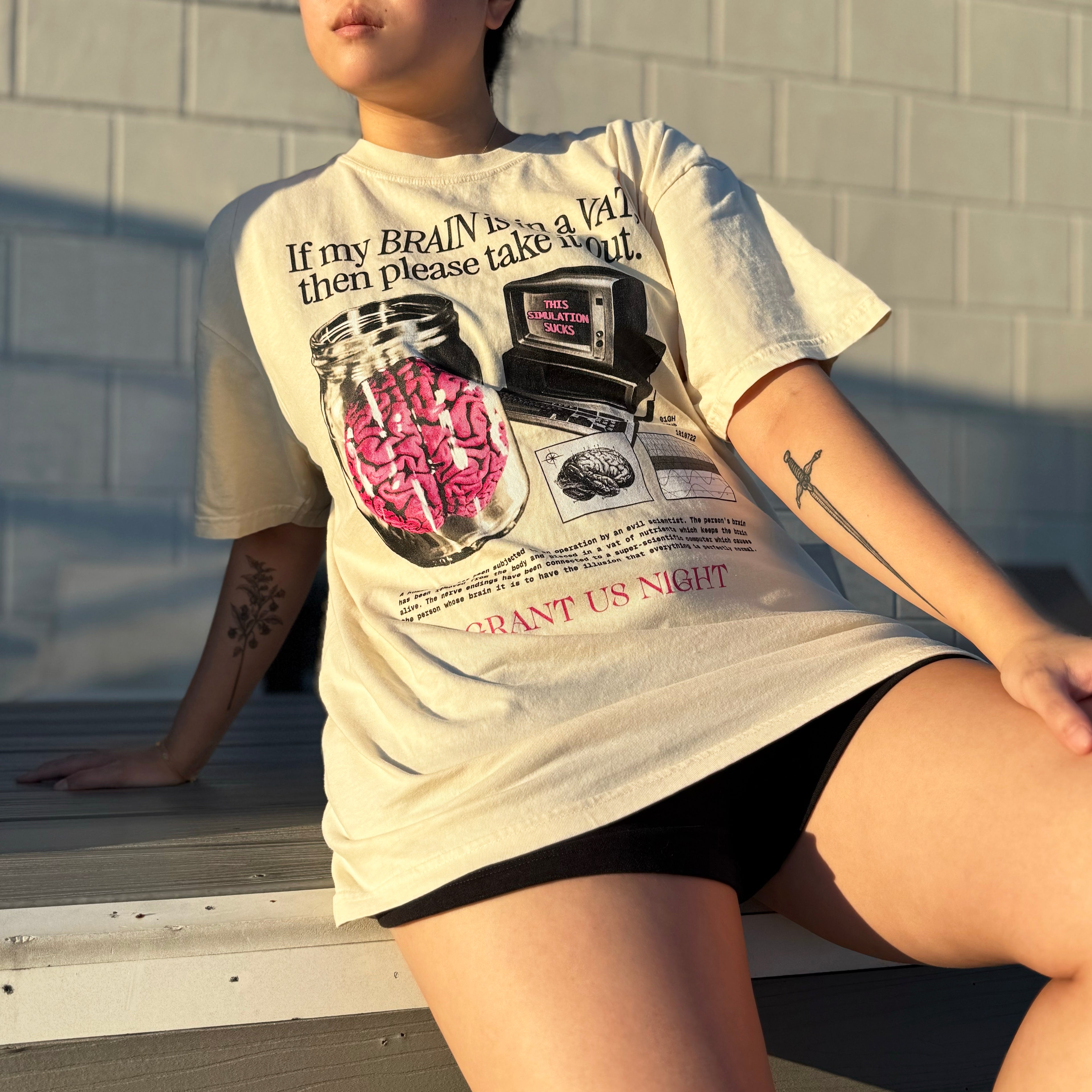 Person wearing a Grant Us Night Vat Brain off white t-shirt with a graphic design of a brain in a jar and an old computer and chart and diagram and text, sitting against a brick wall.