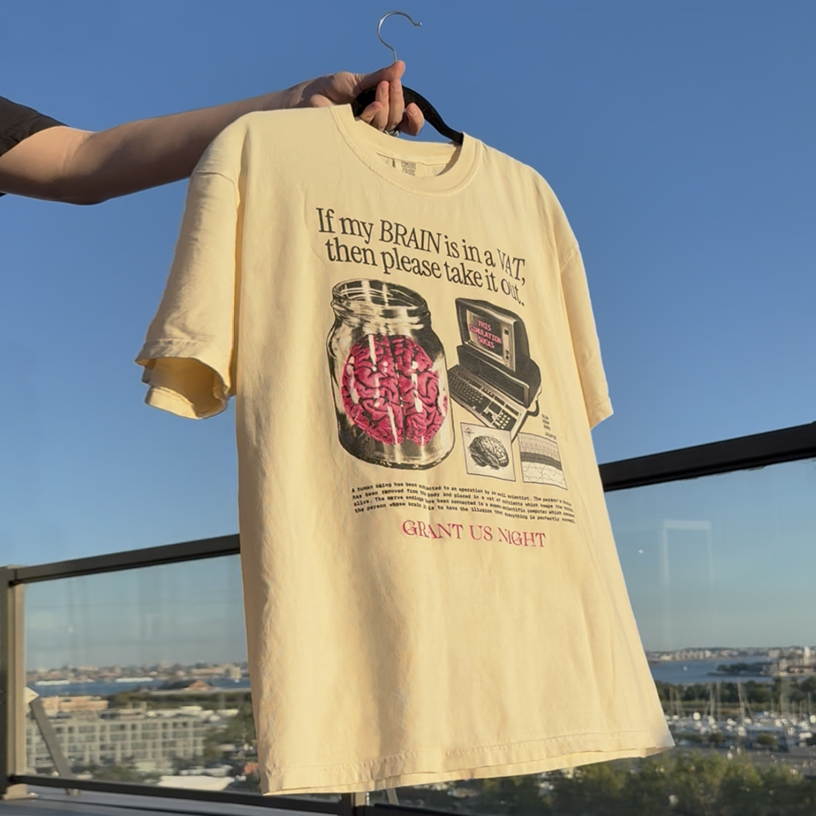 Grant Us Night Vat Brain off white t-shirt with a graphic design of a brain in a jar and an old computer and chart and diagram and text, held on a hanger against a blue sky.